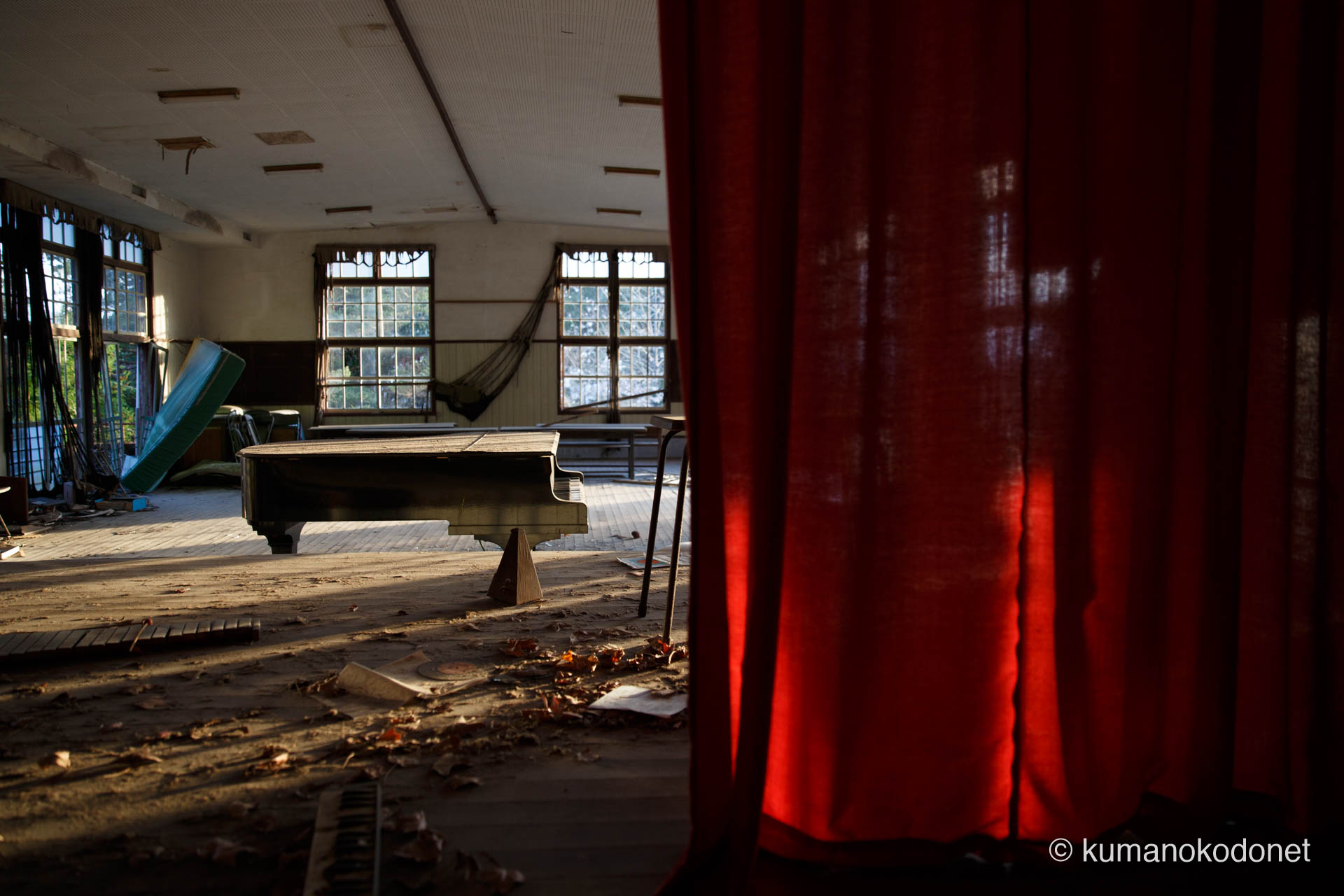 廃校の講堂で、埃を被りながらも佇むピアノを捉えた記録。手前の赤いカーテンが、音の消えた空間に鮮やかな孤独の色彩を添えている。