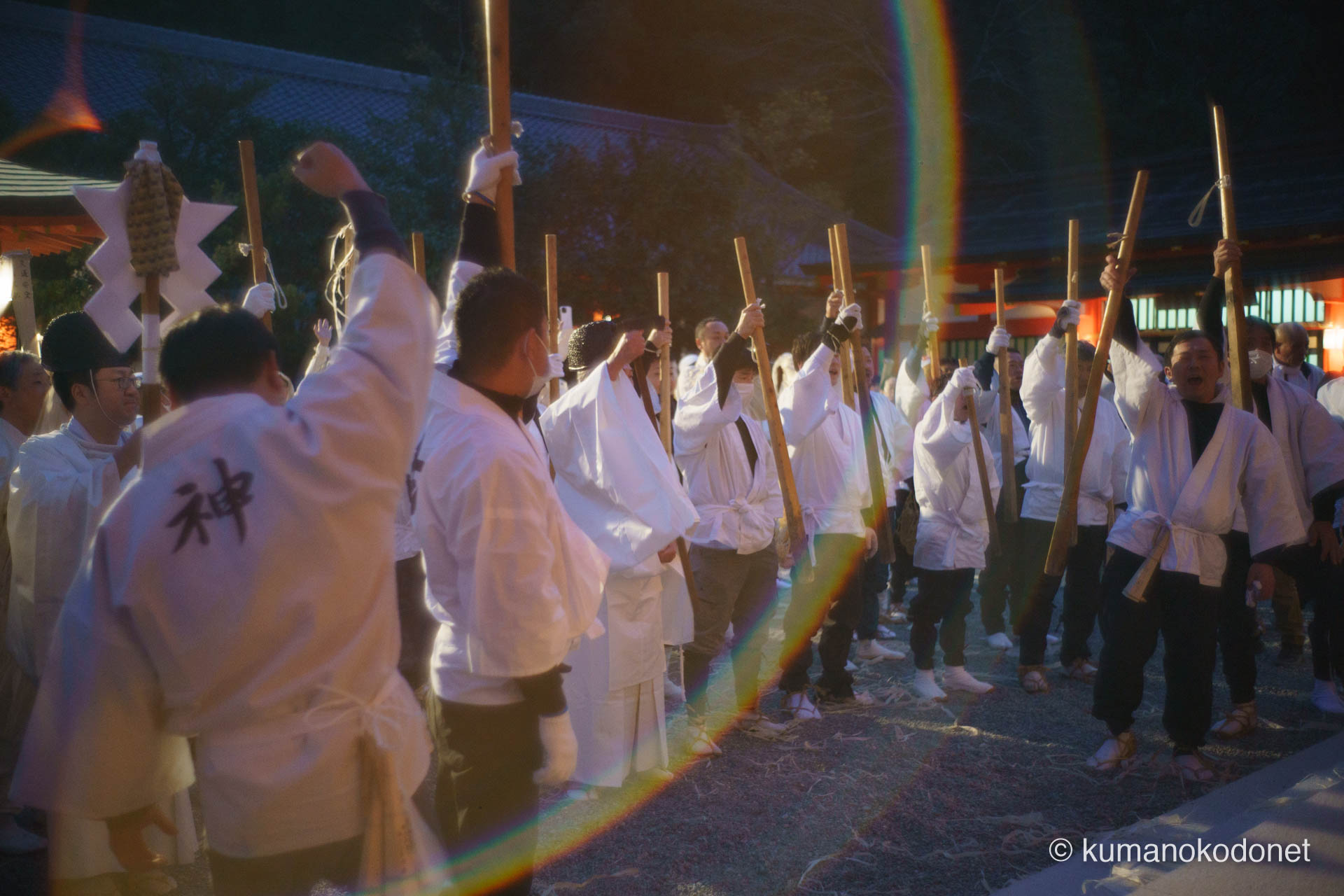 熊野速玉大社の拝殿前、拳を突き上げ咆哮する介釈たちと、静かに祈りを捧げる神職が織りなす極限の情景。画面を横切る虹色の光芒が男たちの決意を祝福するように降り注ぎ、神事の開始を告げる魂の叫びが夜の静寂を震わせる、圧倒的な臨場感に満ちたお燈祭りの様相。 ｜ Kumano Hayatama Taisha, Shingu, Wakayama ｜ SONY α7 ｜ 2026