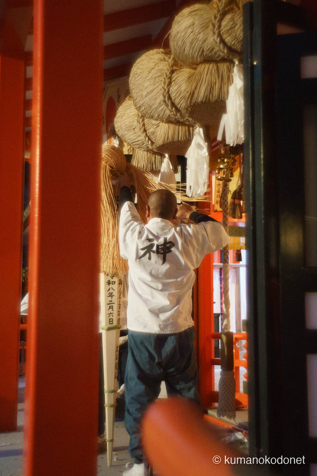 巨大な注連縄が飾られた拝殿で、大松明の先端から白い奉納物を慎重にちぎり取り、賽銭箱の奥へと投げ入れる介釈の後ろ姿。背負った「神」の文字が夕闇に浮かび上がり、祭りの最高潮を前に、聖なる火の魂を神へと捧げる秘儀のような一コマ。 ｜ Kumano Hayatama Taisha, Shingu, Wakayama ｜ SONY α7 ｜ 2026