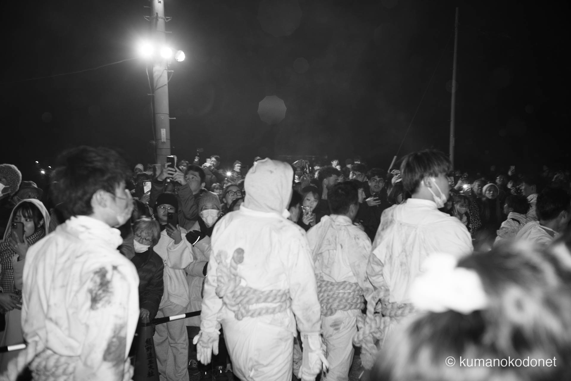 神倉山の麓、街へと溢れ出した上り子たちを迎え入れる、観衆の凄まじい熱気と興奮の渦。白装束の背中の向こう側にひしめき合う無数の視線と、その瞬間を焼き付けようとするスマートフォンの光が交錯し、冬の夜気を一変させるような祝祭ならぬ「神事」の余熱が、モノクロの画面いっぱいに充満する。 ｜ Kamikura, Shingu, Wakayama ｜ SONY α7 ｜ 2026