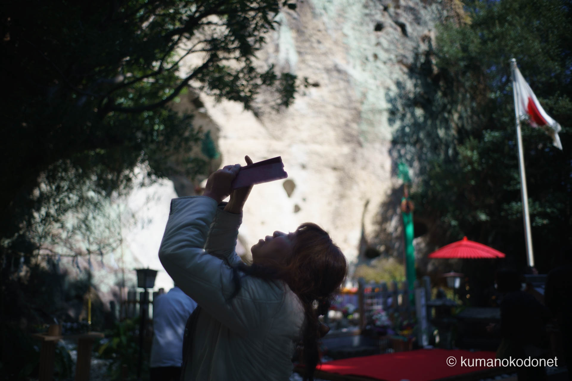 花の窟神社の境内にて、天高く掲げられた三流の幡(みながれのはた)をスマートフォンで撮影する参拝客の後ろ姿。世代を問わず多くの人々が御神体を見上げ、神事の瞬間を記録に収める現代の例大祭の情景。| Hananoiwaya Jinja, Arima, Kumano, Mie | SONY α7 | 2026