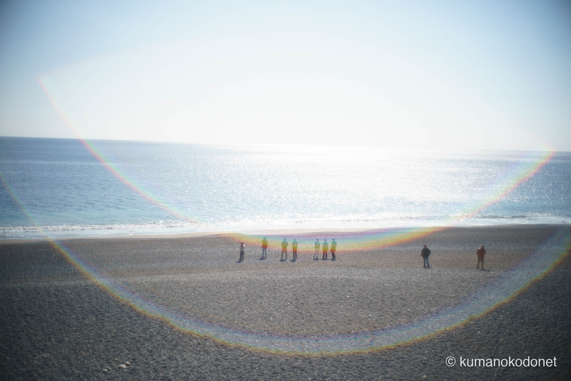 陽光輝く七里御浜(しちりみはま)の波打ち際で、神事を見守る人々の遠景。レンズフレアが描く虹色の輪が浜辺を優しく包み込む、お綱掛け神事の日に現れた奇跡のような一幕。| Shichirimihama Beach, Hananoiwaya Jinja, Arima, Kumano, Mie | SONY α7 | 2026