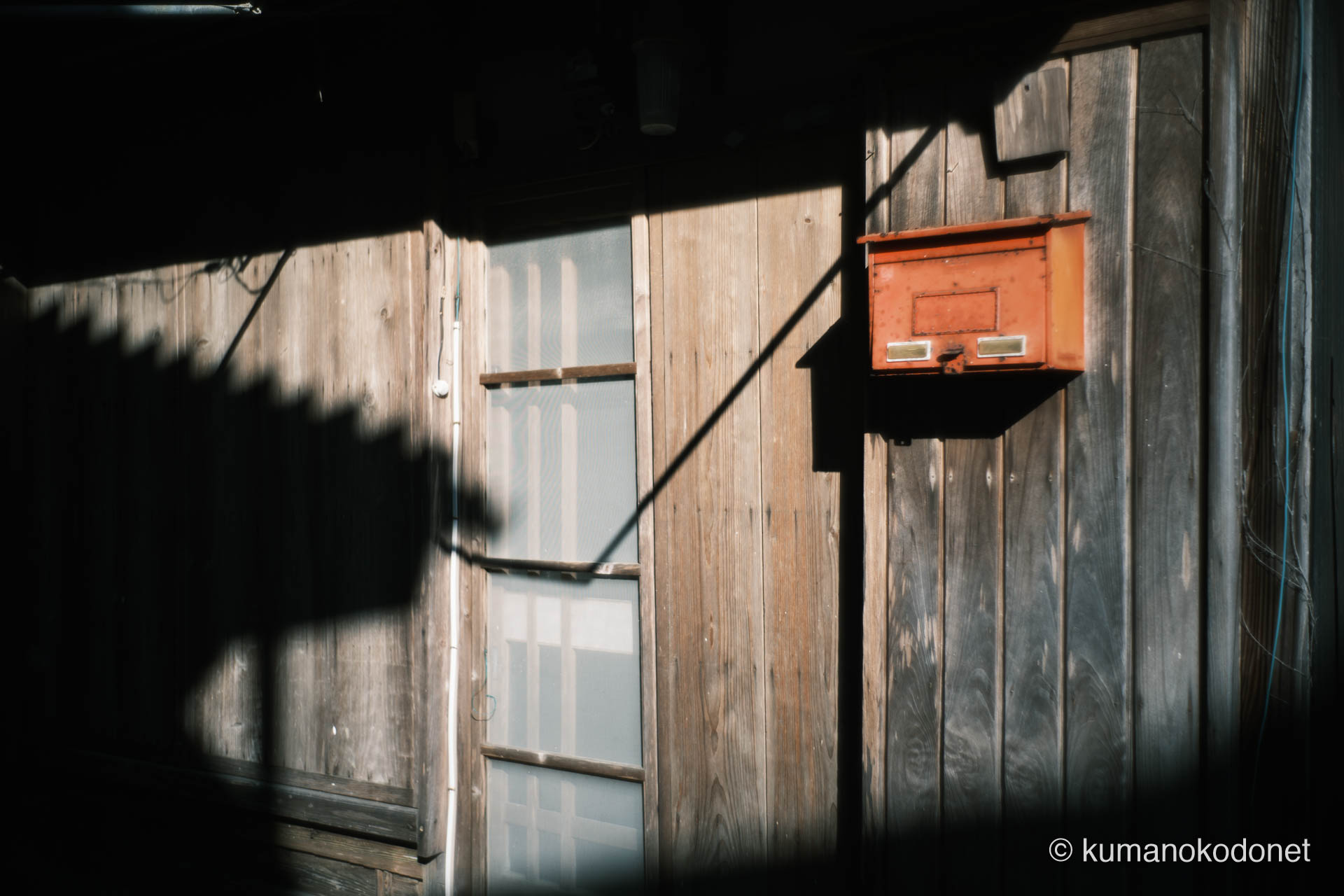 古い木造家屋の壁に掛けられた赤い郵便受け。西日が影を長く落とし、静かな生活の息吹を記録。｜ Kuki, Owase, Mie ｜ FUJIFILM GFX Classic Neg. ｜ 2026