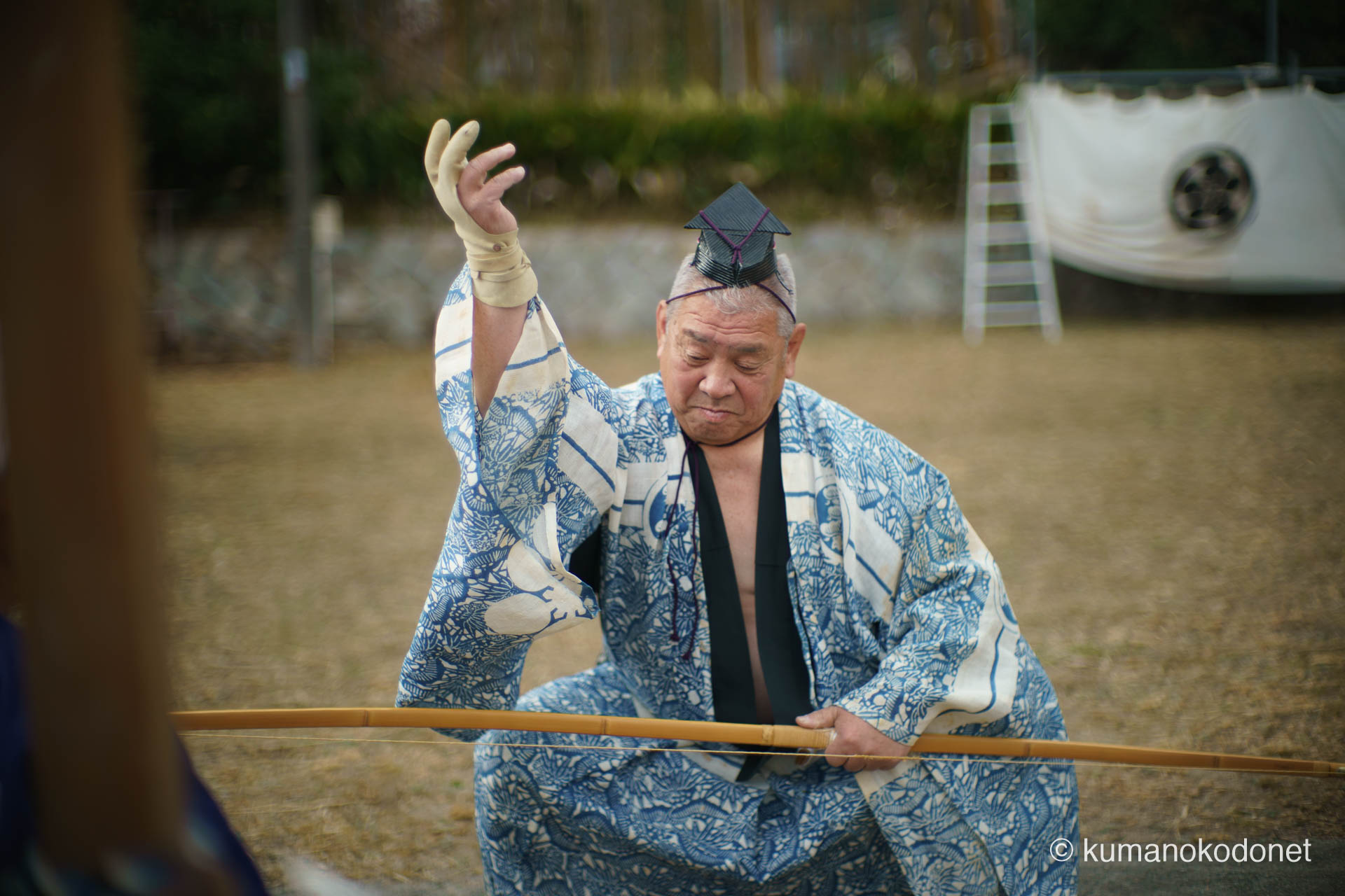 神社そばの空き地で行われる弓行事の合間、傍らに置かれた弓の前に跪き、状態を確認する射手。 一射ごとに繰り返される、道具への敬意と神事への集中を重んじる丁寧な所作を捉えた一枚。空き地の土煙と奉納幟が、祭りのリアリティを物語る。| Tenma Tenjinja, Nachikatsuura, Wakayama | SONY α7 | 2026