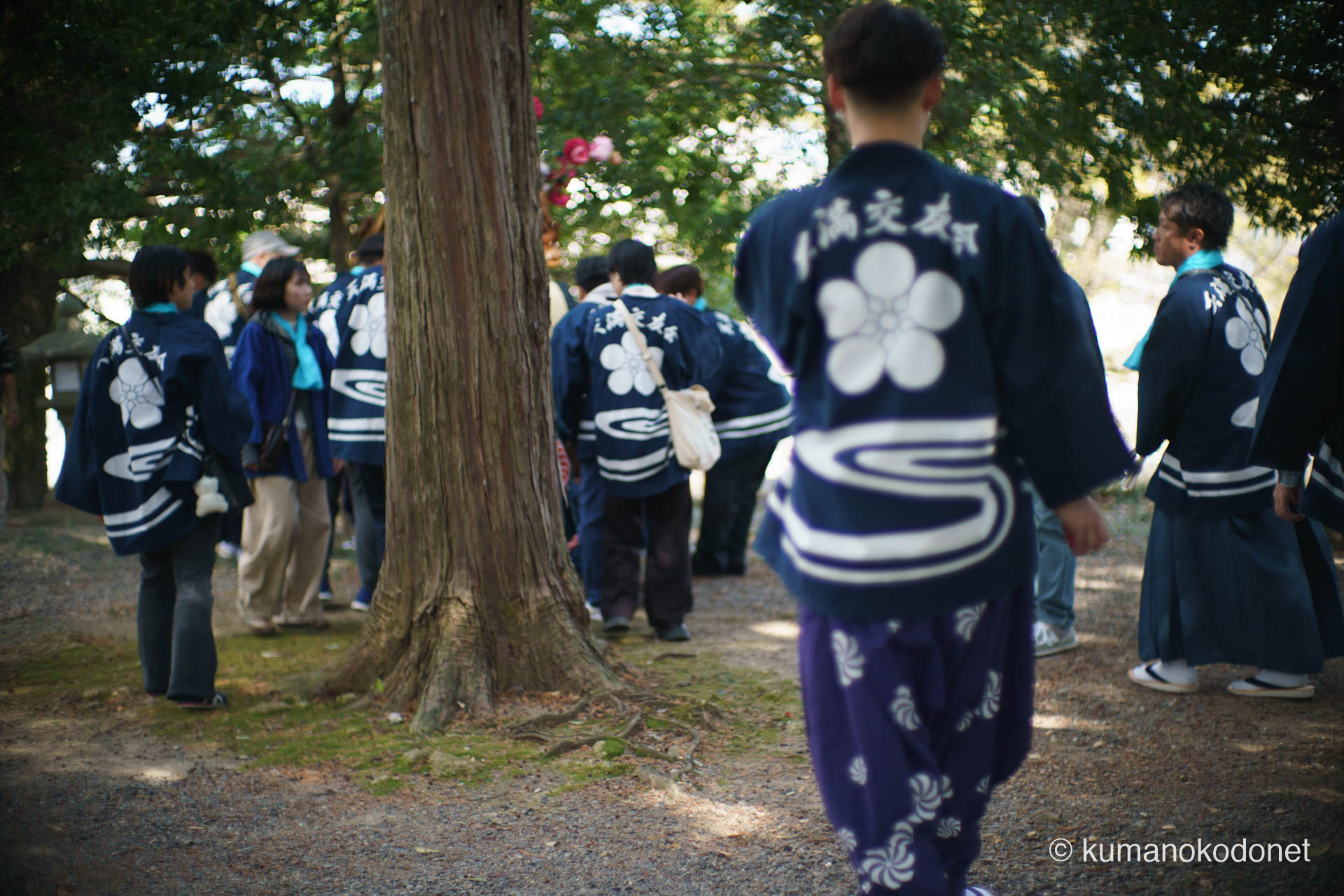 天満天神社の境内で、獅子舞を乗せた山車を囲む天満交友会の男たちの後ろ姿。揃いの法被の背には、地域の伝統と誇りを象徴する紋章が白く浮かび上がる。木漏れ日が降り注ぐ参道を、祭りの熱狂へと向かって歩む彼らの連帯感を捉えた情景。| Tenma Tenjinja, Nachikatsuura, Wakayama | SONY α7 | 2026