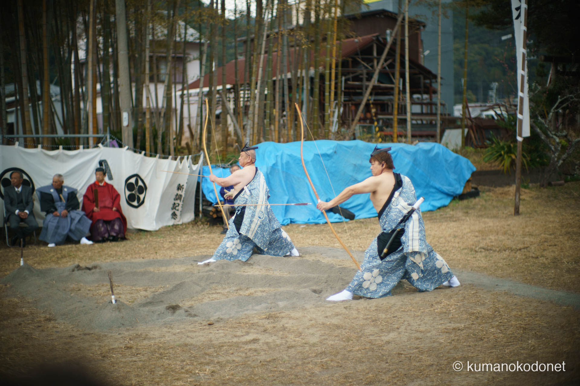 空き地の射場で、深く腰を落として同時に弓を構える二人の射手。背景の奉納幟が風に揺れる中、生活の場が極限の緊張感に包まれる、弓行事クライマックスの躍動感溢れる一景。| Tenma Tenjinja, Nachikatsuura, Wakayama | SONY α7 | 2026