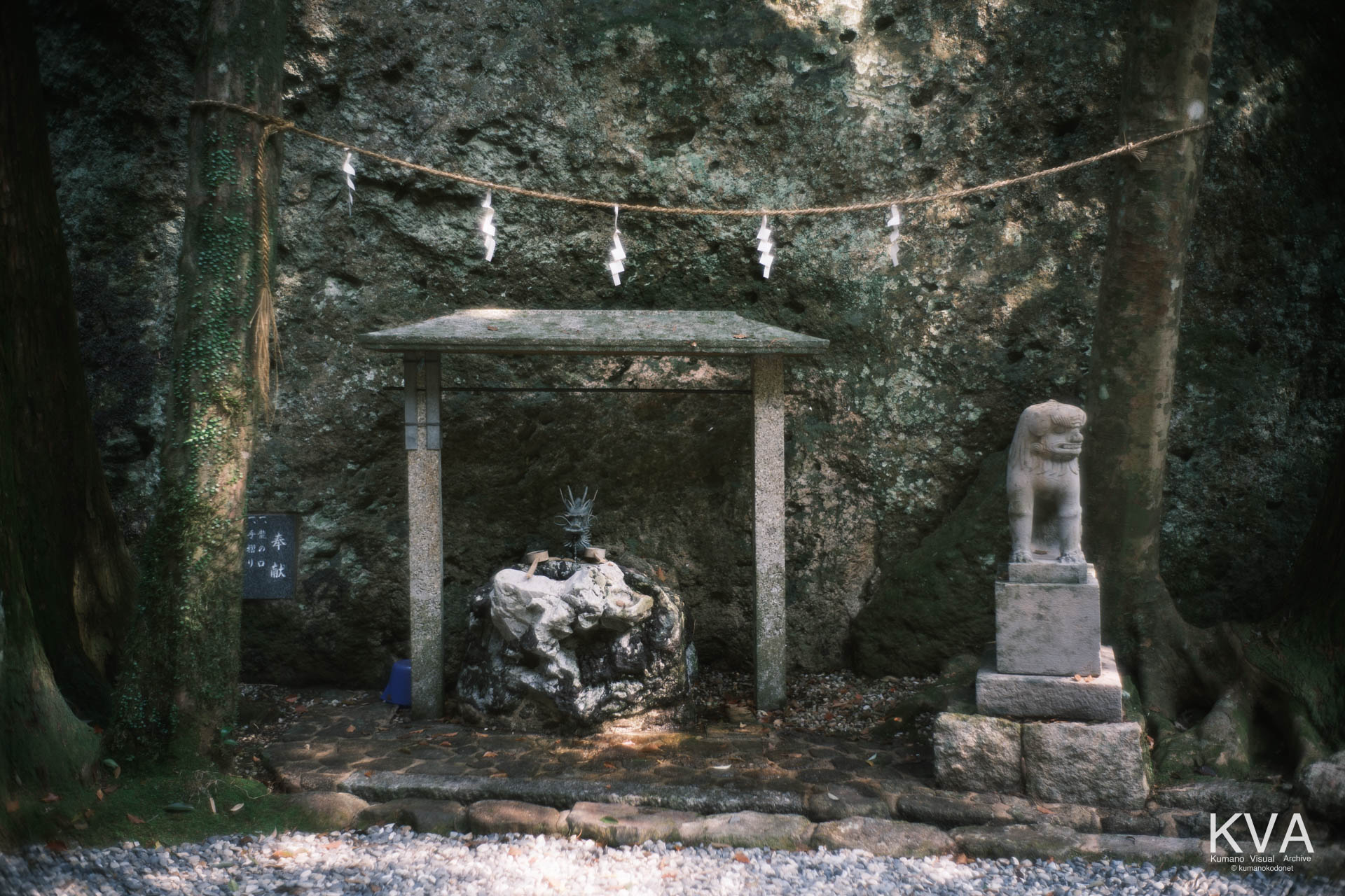 巨大な岩窟を背に鎮座する手水舎（てみずや）と、傍らに立つ狛犬および奉献銘板の全景