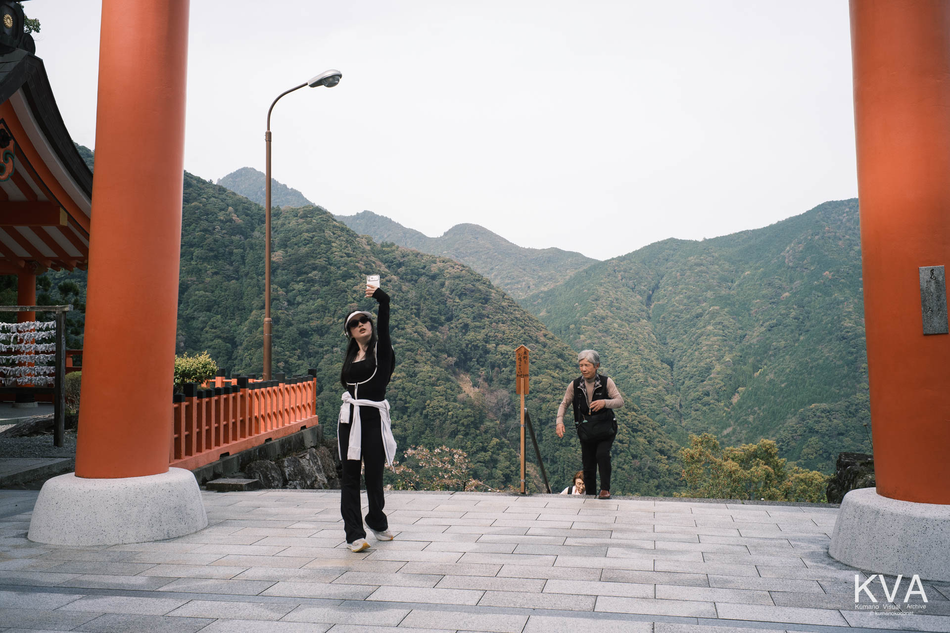 大鳥居の間でスマートフォン自撮りをする外国人女性と、石段を登る高齢の参拝者
