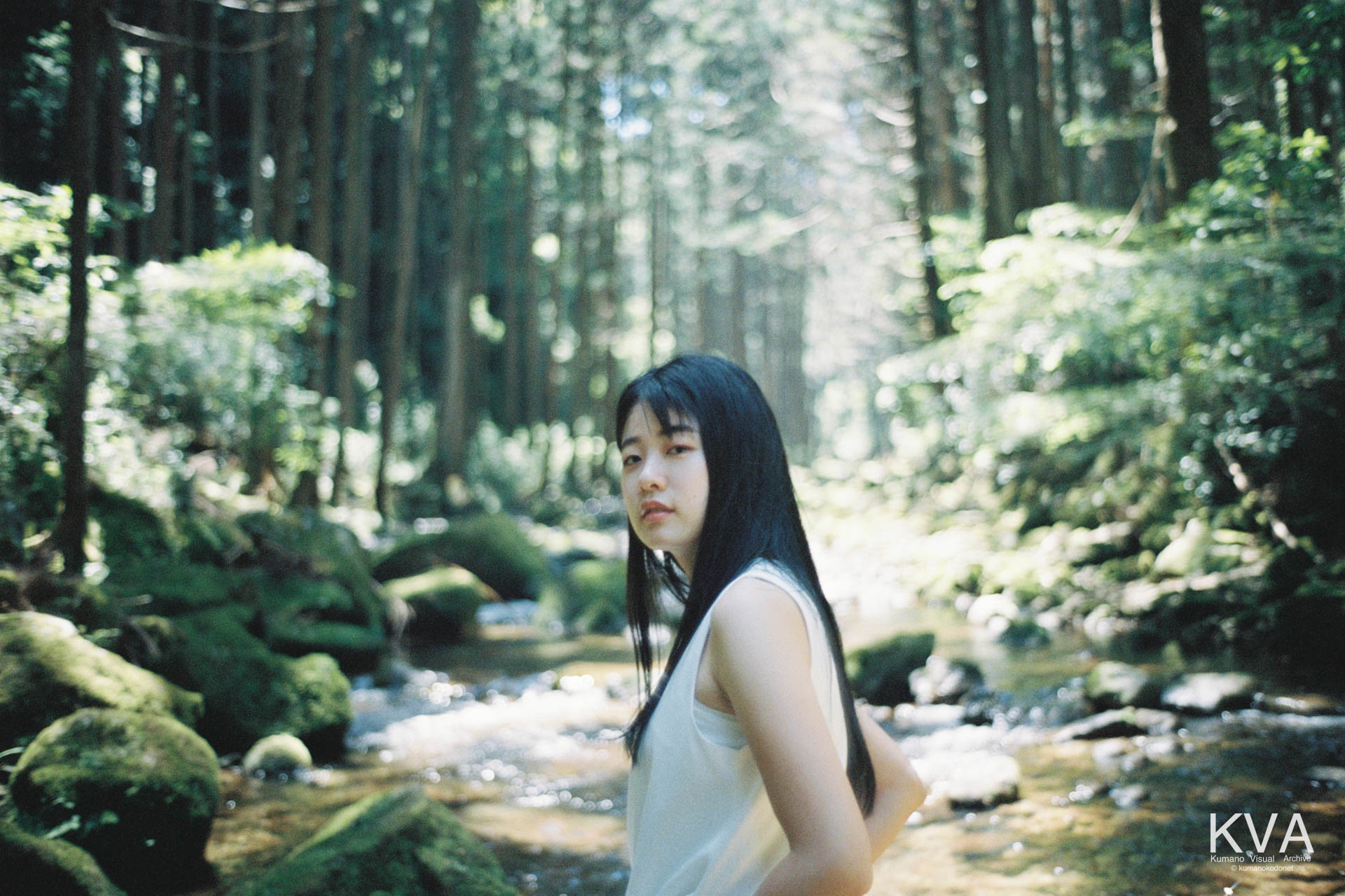 清流と緑に囲まれた河原を背景に、ふと振り返ってカメラに視線を向ける女性のポートレート
