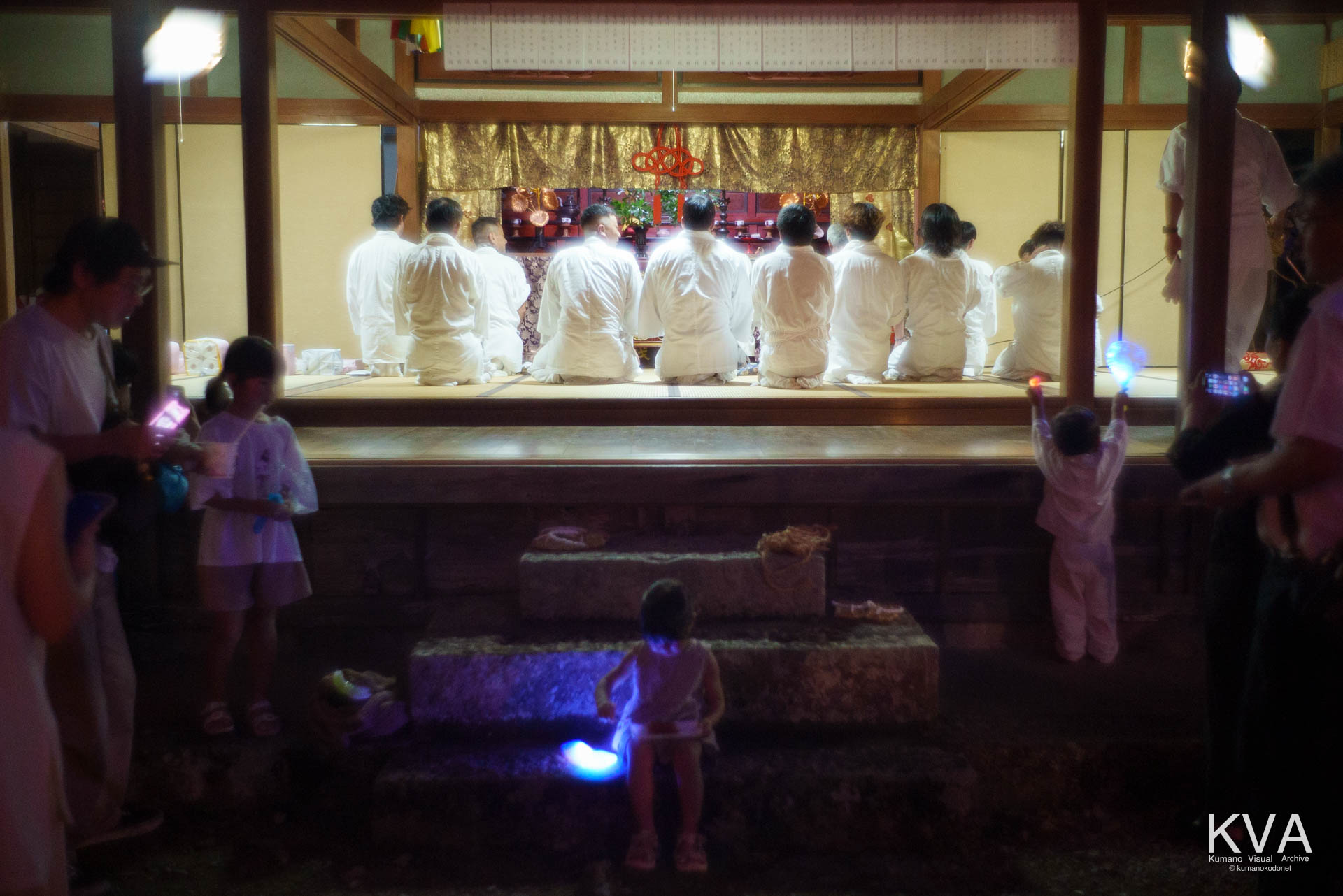 本堂で黙祷を捧げる白装束の男たちと境内に佇む子どもたち